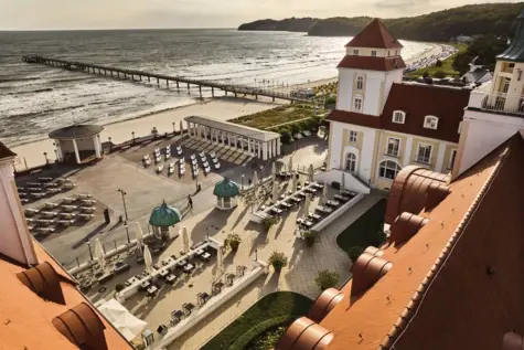 Das Kurhaus Binz aus der Vogelperspektive mit seinen weißen Fassaden und roten Dächern. Aus der Luft ist über das Kurhaus hinweg auf dessen Innenhof mit Sitzmöglichkeiten und den angrenzenden Strand mit Meer zu blicken. 