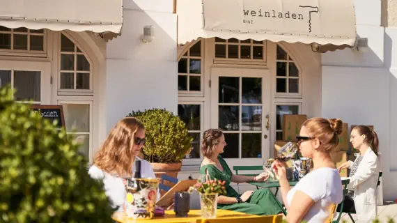 Ein Weinladen mit Sitzmöglichkeiten von außen. An bunten Tischen sitzen Menschen, genießen ihren Wein und die warme Sonne. 