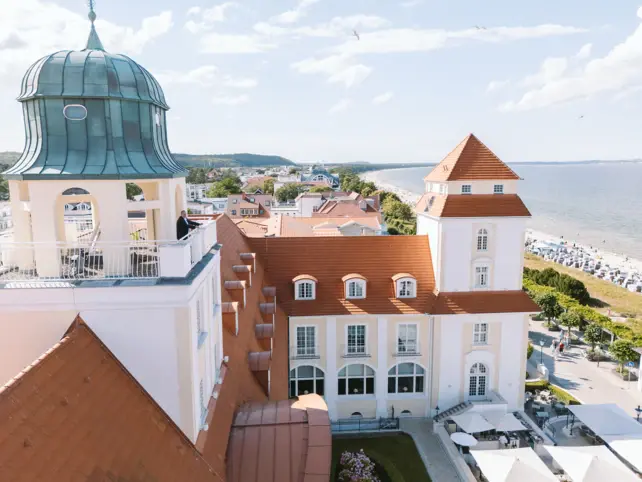 Eine Vorderansicht des hellen Kurhauses binz mit vielen Fenstern und einem roten Dach. 