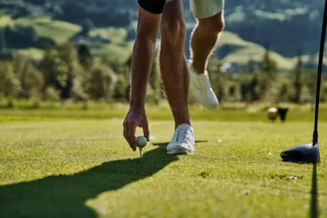 Person positioniert einen Golfball auf einem Rasen vor einer Bergkulisse mit grünen Bäumen im Hintergrund..